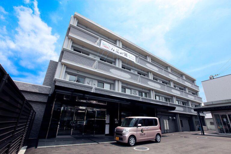 介護施設の建物外観。複数階建てで駐車場に送迎車が停まっている。