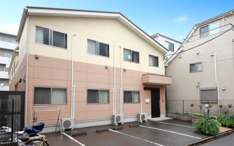 2階建ての介護施設の建物外観。クリーム色と桜色の外壁、複数の窓が配置されている。